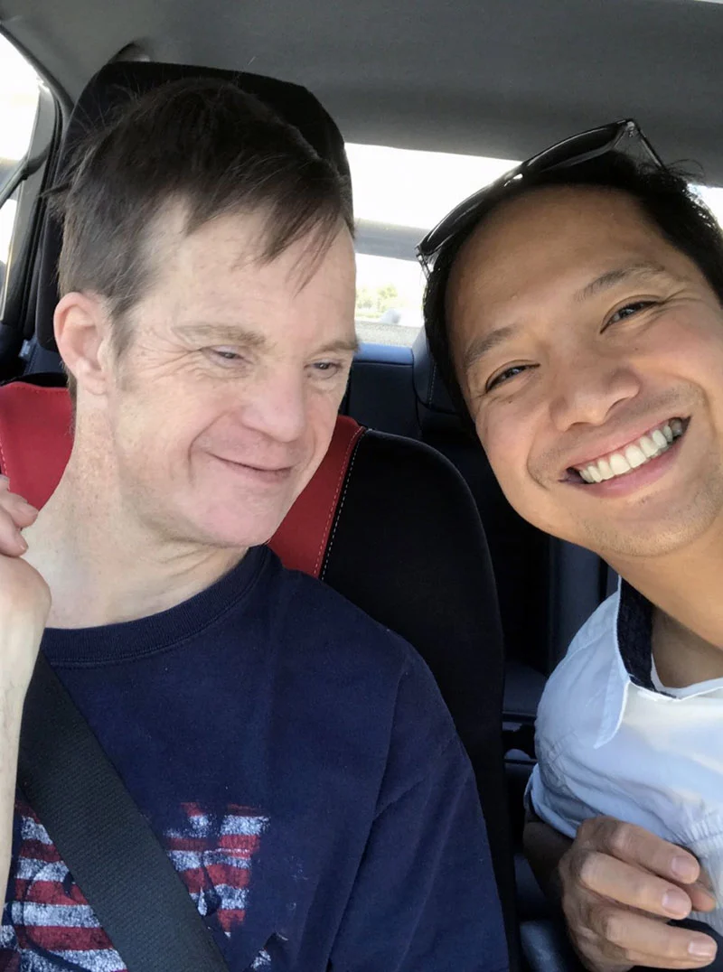 Two smiling men sit in a car; one has Down syndrome. Their joyful expressions embody the Christopher Smith Foundation’s mission of caregiver support and disability advocacy, highlighting the bond between caregivers and those they support.| A Non Profit For Caregivers | Christopher Smith Foundation