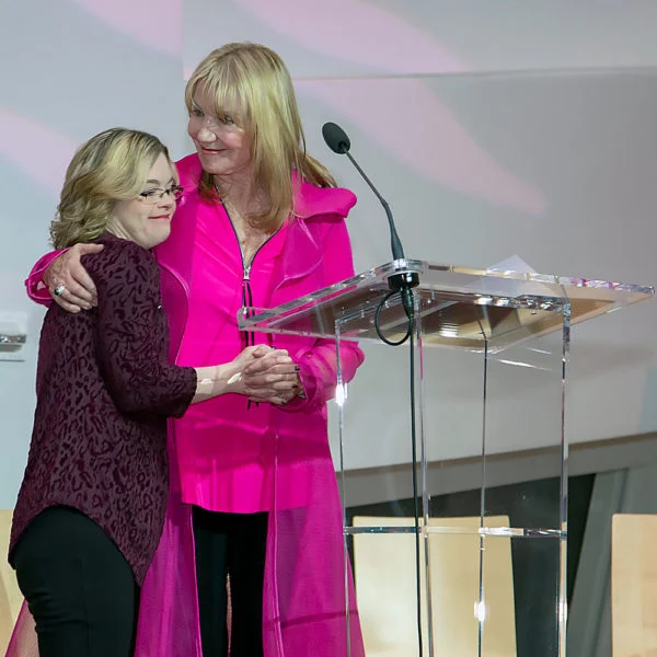 A woman in a bright pink outfit embraces a younger woman with glasses on stage beside a podium, symbolizing caregiver support at a Christopher Smith Foundation event celebrating caregivers of people with disabilities.| A Non Profit For Caregivers | Christopher Smith Foundation