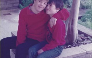 Two boys in red sweaters sit together on a stone bench, one with his arm around the other, smiling warmly. The scene reflects the Christopher…| A Non Profit For Caregivers | Christopher Smith Foundation