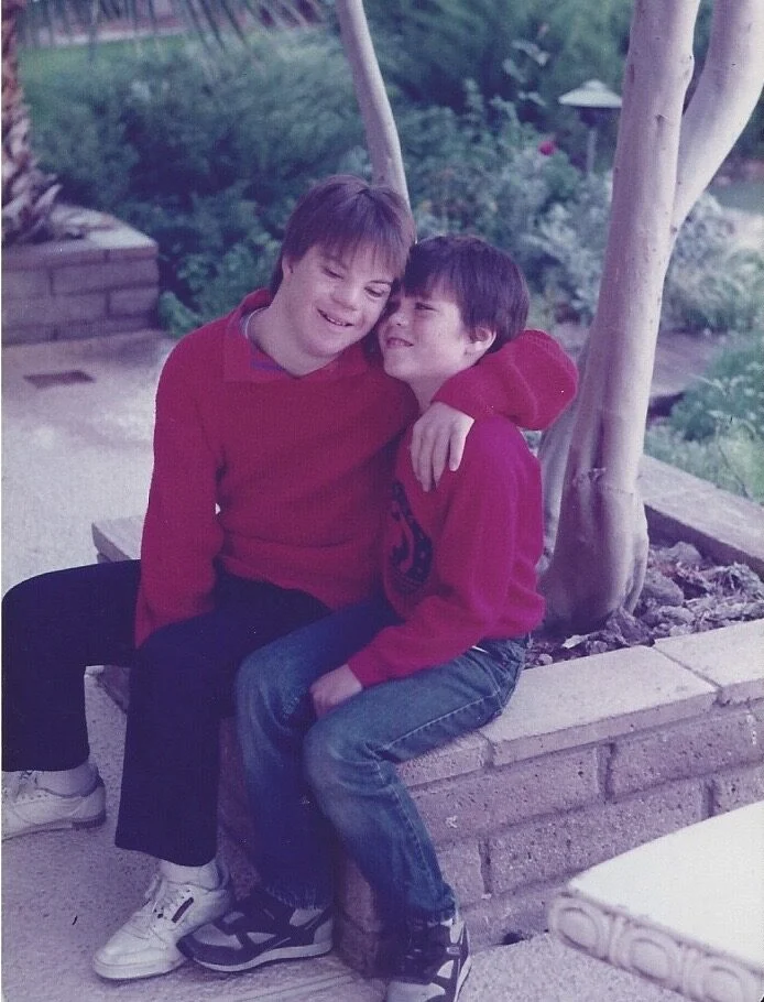 Two boys in red sweaters sit together on a stone bench, one with his arm around the other, smiling warmly. The scene reflects the Christopher…| A Non Profit For Caregivers | Christopher Smith Foundation