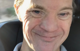 A man with light skin and short hair smiles warmly while seated in a car, wearing a light blue polo shirt. The image reflects the Christopher Smith Foundation's mission of caregiver support and disability advocacy.| A Non Profit For Caregivers | Christopher Smith Foundation