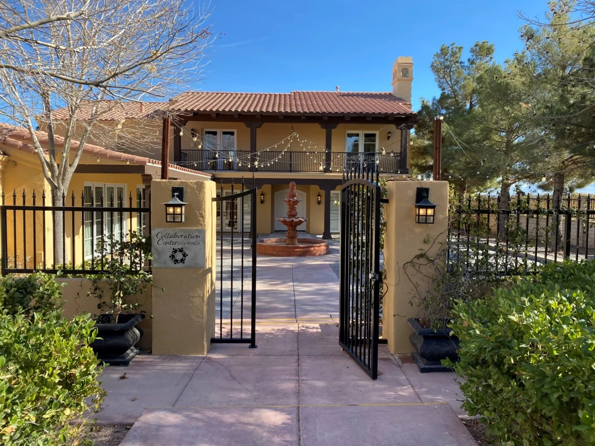 A sunny courtyard with a welcoming open gate leads to a beige building with a red-tiled roof, surrounded by trees. The Christopher Smith Foundation promotes caregiver support and disability advocacy in this serene setting.| A Non Profit For Caregivers | Christopher Smith Foundation