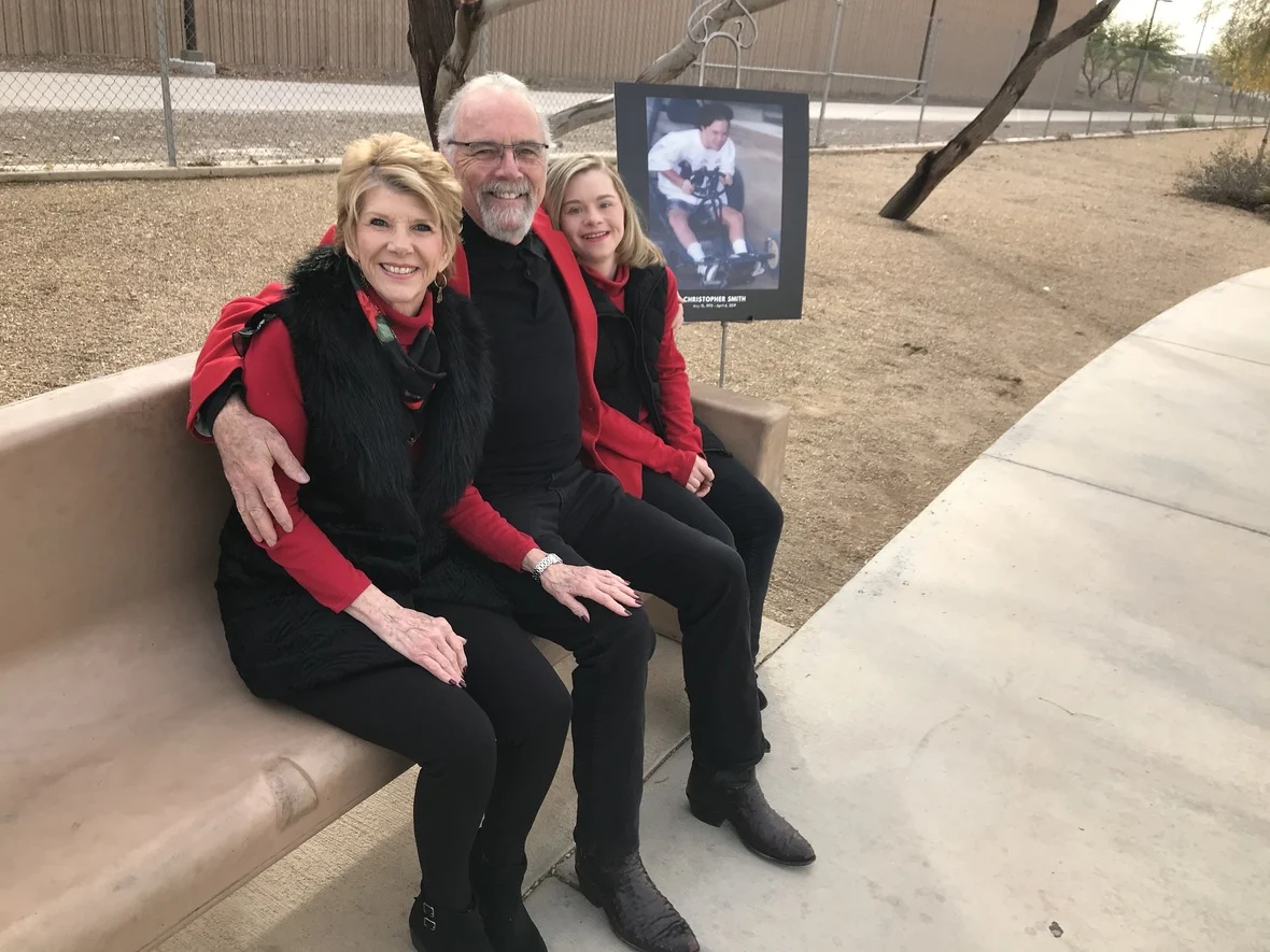 A family sits on a park bench smiling, with a photo of a person in a wheelchair beside them, symbolizing caregiver support by the Christopher Smith Foundation.| A Non Profit For Caregivers | Christopher Smith Foundation
