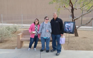 A group of three people stands outdoors by a bench, holding gifts. A framed photo on an easel is beside them. This image highlights caregiver recognition by the Christopher Smith Foundation, supporting caregivers of people with disabilities.| A Non Profit For Caregivers | Christopher Smith Foundation