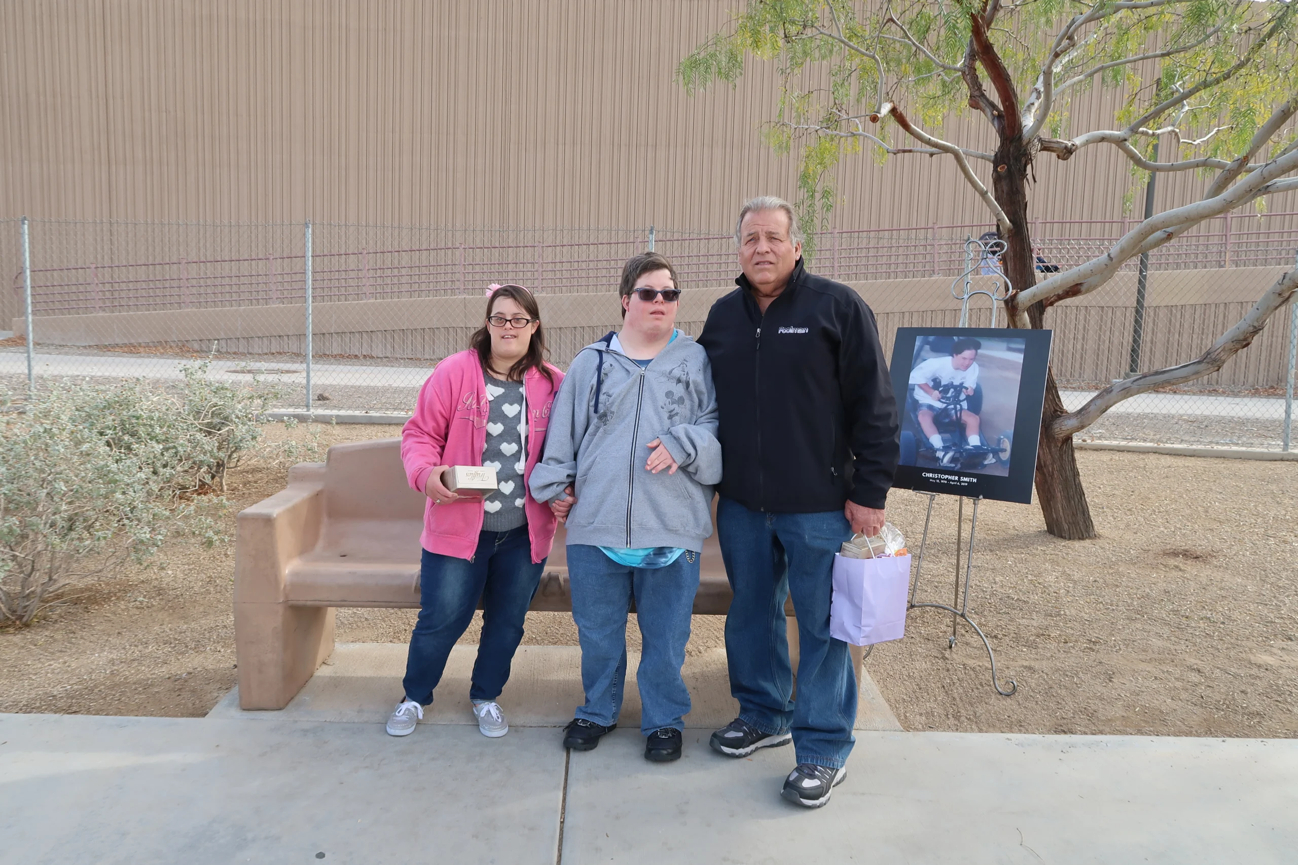 A group of three people stands outdoors by a bench, holding gifts. A framed photo on an easel is beside them. This image highlights caregiver recognition by the Christopher Smith Foundation, supporting caregivers of people with disabilities.| A Non Profit For Caregivers | Christopher Smith Foundation