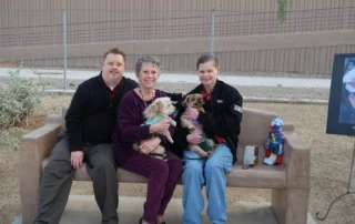 A woman sits on a bench between two men, each holding a small dog, smiling in a park setting. The Christopher Smith Foundation supports caregivers of people with disabilities, fostering community and compassion.| A Non Profit For Caregivers | Christopher Smith Foundation