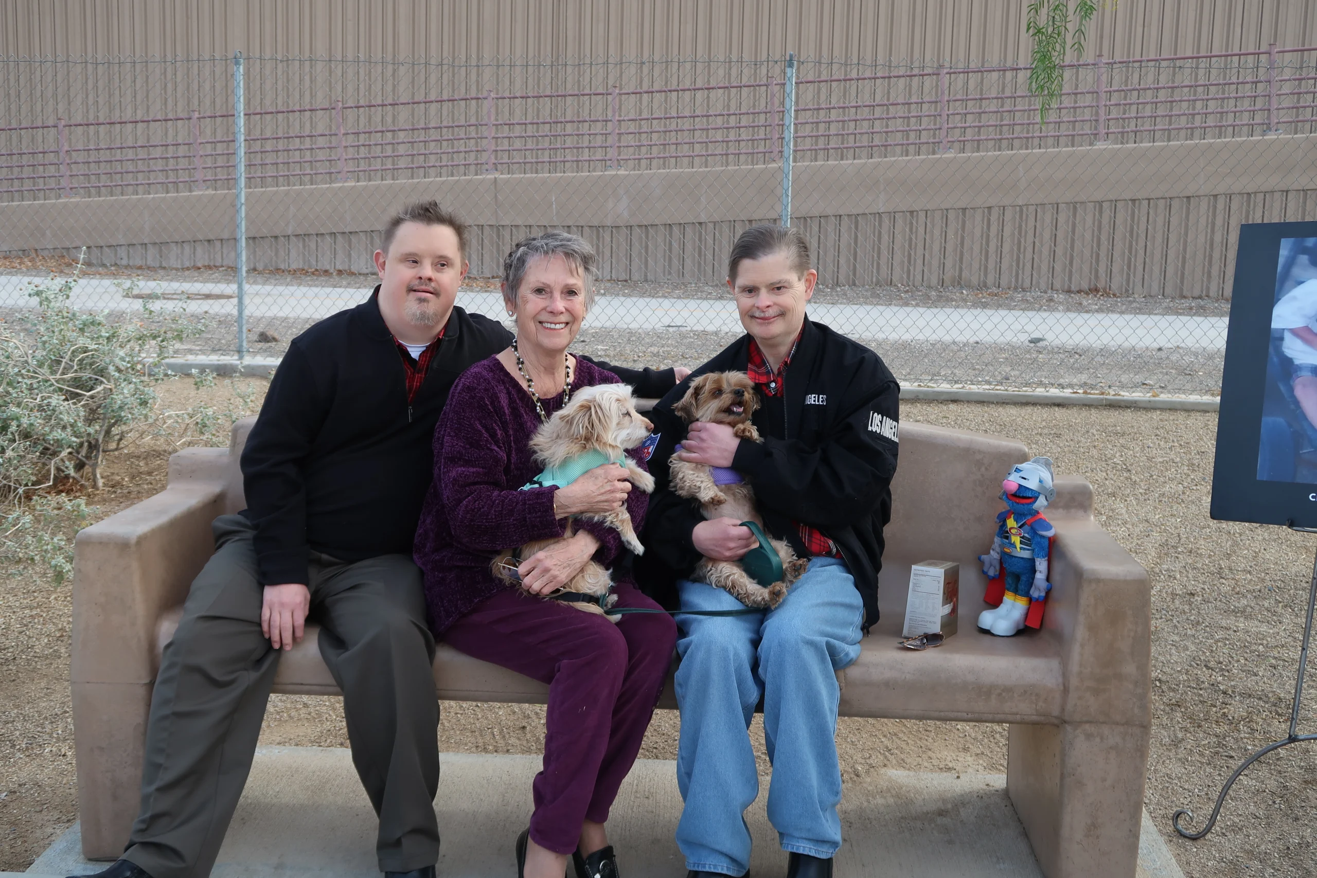A woman sits on a bench between two men, each holding a small dog, smiling in a park setting. The Christopher Smith Foundation supports caregivers of people with disabilities, fostering community and compassion.| A Non Profit For Caregivers | Christopher Smith Foundation