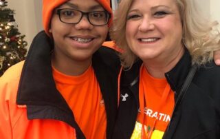 Two people in orange jackets and hats smile warmly, standing close together indoors. A Christmas tree is in the background. This image embodies the Christopher…| A Non Profit For Caregivers | Christopher Smith Foundation