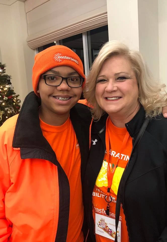 Two people in orange jackets and hats smile warmly, standing close together indoors. A Christmas tree is in the background. This image embodies the Christopher…| A Non Profit For Caregivers | Christopher Smith Foundation