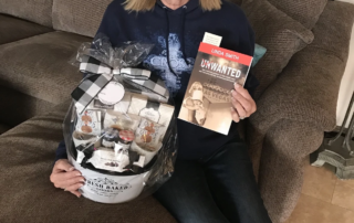 A smiling woman sits on a brown couch holding a gift basket and a book. This image represents caregiver recognition by the Christopher Smith Foundation, highlighting the importance of supporting caregivers of people with disabilities.| A Non Profit For Caregivers | Christopher Smith Foundation