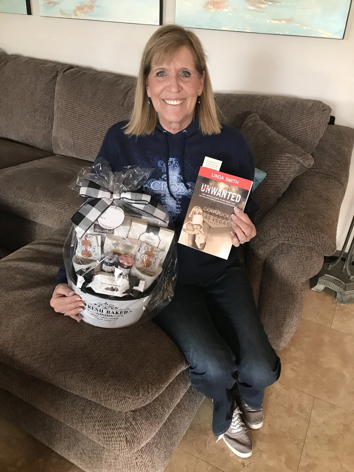 A smiling woman sits on a brown couch holding a gift basket and a book. This image represents caregiver recognition by the Christopher Smith Foundation, highlighting the importance of supporting caregivers of people with disabilities.| A Non Profit For Caregivers | Christopher Smith Foundation