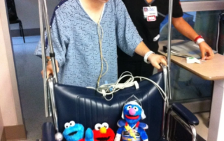 A caregiver supports a man in a hospital gown using a walker, with three plush toys on a wheelchair nearby, representing the Christopher Smith Foundation's mission of caregiver support and disability advocacy.| A Non Profit For Caregivers | Christopher Smith Foundation