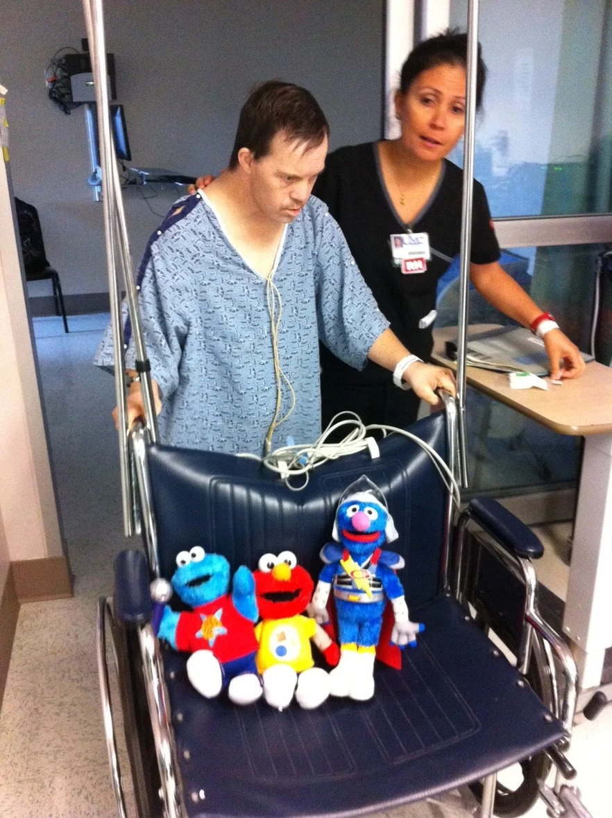 A caregiver supports a man in a hospital gown using a walker, with three plush toys on a wheelchair nearby, representing the Christopher Smith Foundation's mission of caregiver support and disability advocacy.| A Non Profit For Caregivers | Christopher Smith Foundation