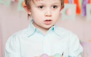 A young child in a light blue shirt holds a carton with pastel eggs, standing against a colorful, blurred background. This image represents the joy…| A Non Profit For Caregivers | Christopher Smith Foundation
