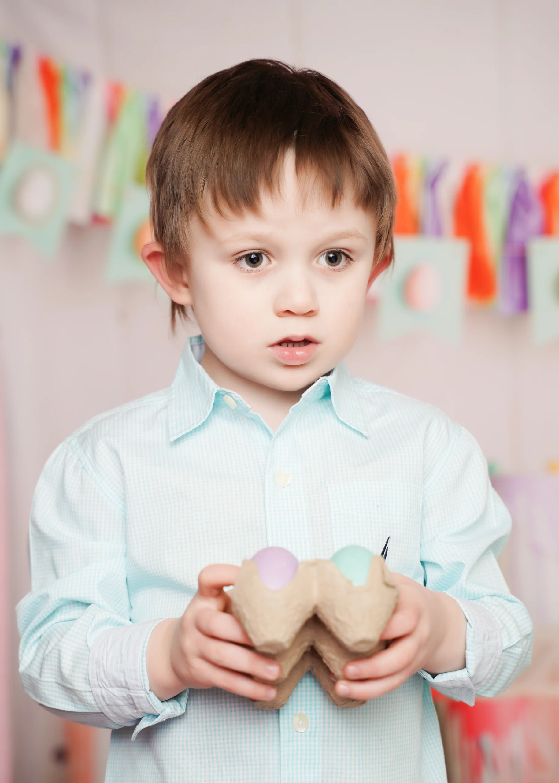 A young child in a light blue shirt holds a carton with pastel eggs, standing against a colorful, blurred background. This image represents the joy…| A Non Profit For Caregivers | Christopher Smith Foundation
