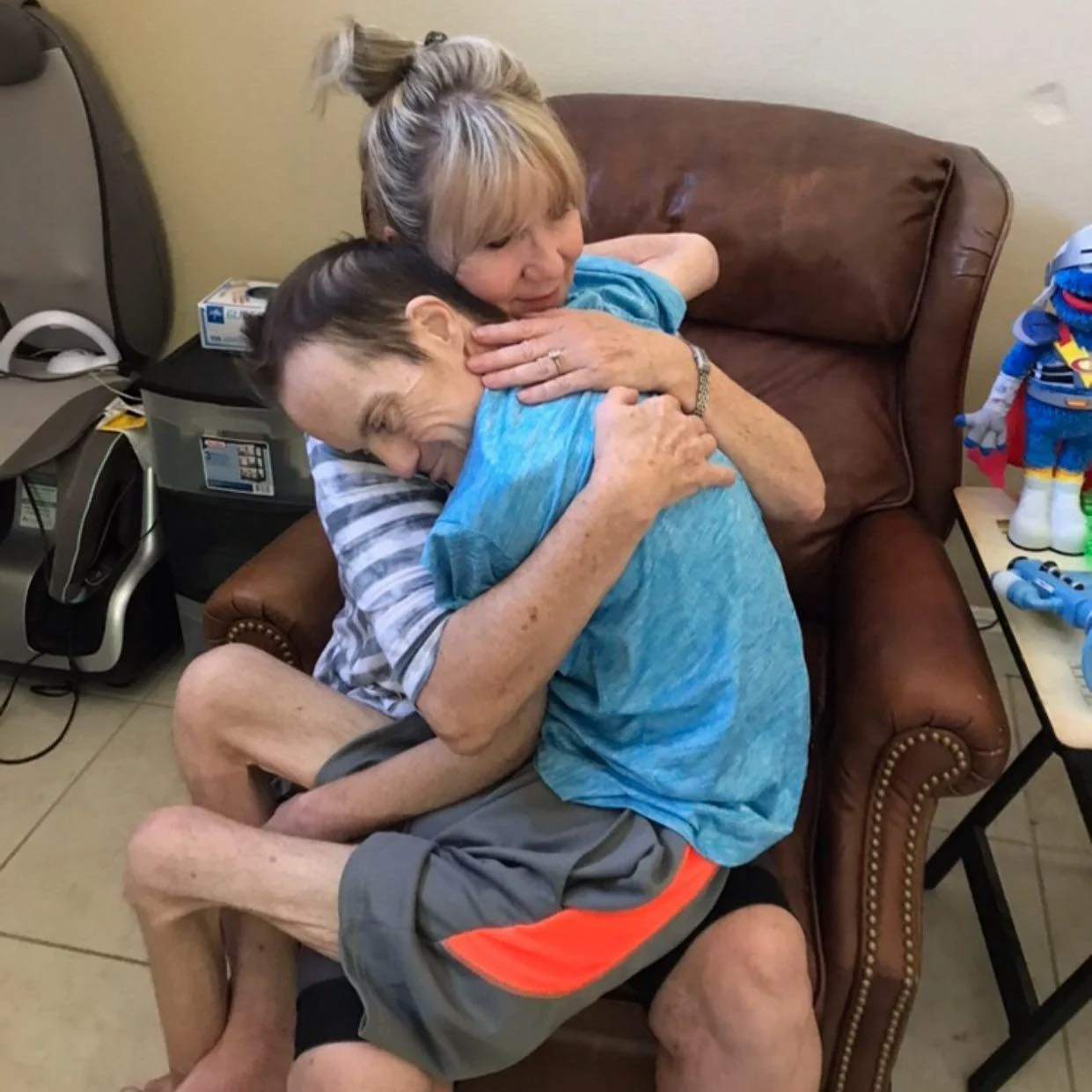 A woman warmly embraces a person with a disability seated on her lap in a cozy living room, embodying the caregiving support celebrated by the Christopher Smith Foundation. A colorful toy robot sits nearby, adding a sense of joy and community.| A Non Profit For Caregivers | Christopher Smith Foundation