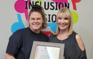 Two women smiling and holding a framed certificate in front of a colorful "Inclusion" sign. The certificate recognizes caregiver support from the Christopher Smith Foundation, celebrating contributions to the disability community.| A Non Profit For Caregivers | Christopher Smith Foundation