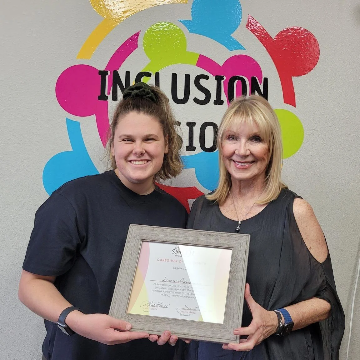 Two women smiling and holding a framed certificate in front of a colorful "Inclusion" sign. The certificate recognizes caregiver support from the Christopher Smith Foundation, celebrating contributions to the disability community.| A Non Profit For Caregivers | Christopher Smith Foundation