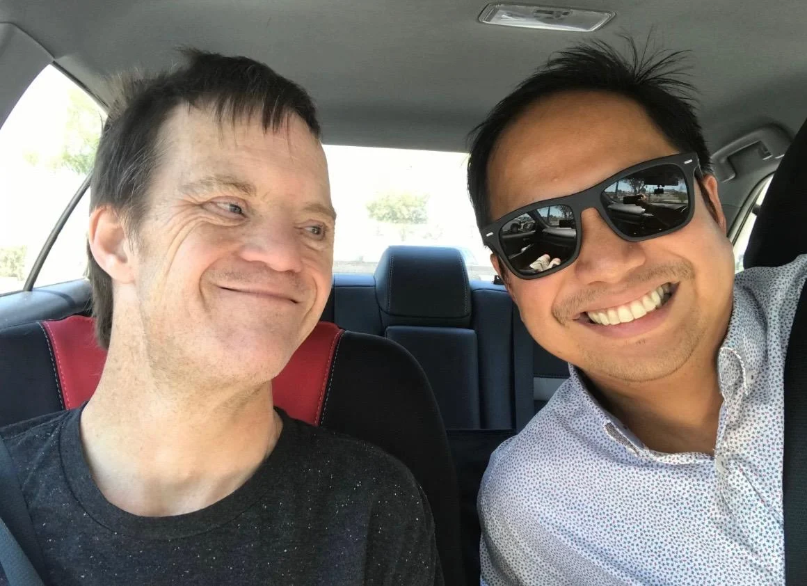 A man wearing sunglasses smiles warmly at another man in a car. They share a joyful moment, highlighting caregiver support within the disability community, reflecting the mission of the Christopher Smith Foundation.| A Non Profit For Caregivers | Christopher Smith Foundation