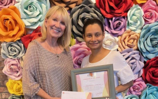Two women stand smiling in front of a colorful backdrop of large paper flowers. One woman holds a "Caregiver of the Month" certificate from the Christopher Smith Foundation, highlighting caregiver recognition and support.| A Non Profit For Caregivers | Christopher Smith Foundation