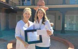 Two women smiling, one holding a certificate and the other an envelope, stand in front of a fountain. They are being recognized by the Christopher Smith Foundation for their caregiver support in the disability community.| A Non Profit For Caregivers | Christopher Smith Foundation