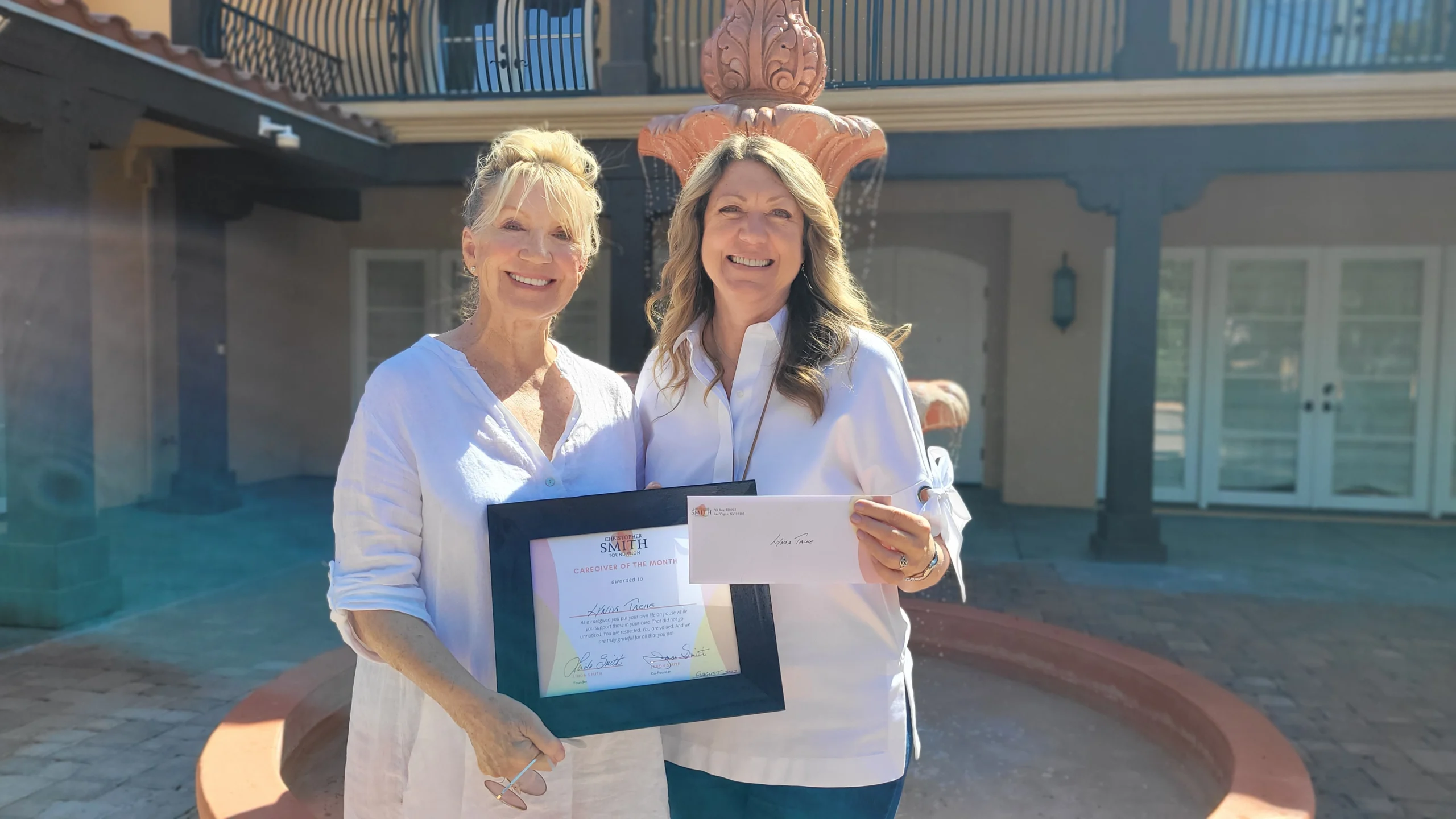 Two women smiling, one holding a certificate and the other an envelope, stand in front of a fountain. They are being recognized by the Christopher Smith Foundation for their caregiver support in the disability community.| A Non Profit For Caregivers | Christopher Smith Foundation