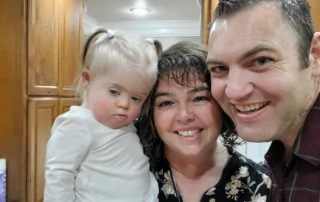 A smiling family with a young child with Down syndrome is embraced by two adults, highlighting family caregivers. The Christopher Smith Foundation supports caregivers of people with disabilities through recognition and advocacy.| A Non Profit For Caregivers | Christopher Smith Foundation