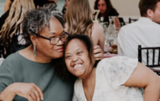 A woman with gray hair and glasses embraces a young woman with a joyful smile at a gathering, symbolizing the Christopher Smith Foundation's commitment to caregiver support and disability advocacy in the community.| A Non Profit For Caregivers | Christopher Smith Foundation
