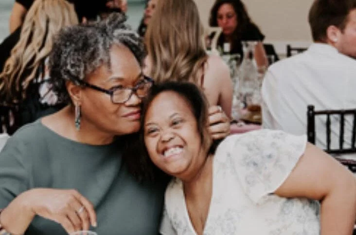 A woman with gray hair and glasses embraces a young woman with a joyful smile at a gathering, symbolizing the Christopher Smith Foundation's commitment to caregiver support and disability advocacy in the community.| A Non Profit For Caregivers | Christopher Smith Foundation