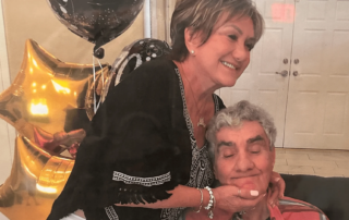 A smiling woman gently holds the face of an elderly man sitting in a chair, surrounded by gold star balloons. This touching moment highlights caregiver support as celebrated by the Christopher Smith Foundation.| A Non Profit For Caregivers | Christopher Smith Foundation