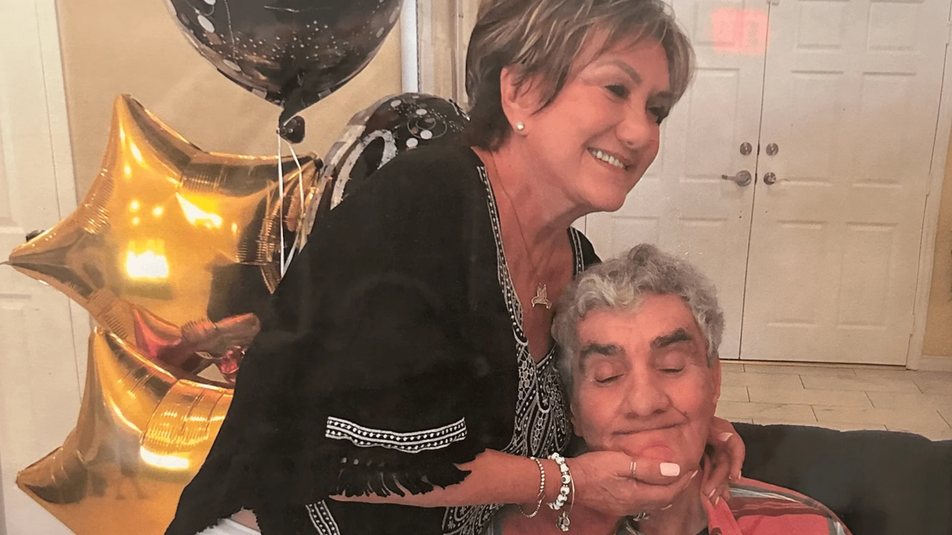 A smiling woman gently holds the face of an elderly man sitting in a chair, surrounded by gold star balloons. This touching moment highlights caregiver support as celebrated by the Christopher Smith Foundation.| A Non Profit For Caregivers | Christopher Smith Foundation