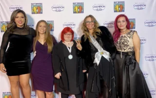 Five women stand smiling in elegant dresses at a Christopher Smith Foundation event, celebrating caregiver recognition. The backdrop features logos and the phrase "Champion of the Year," highlighting support for the disability community.| A Non Profit For Caregivers | Christopher Smith Foundation