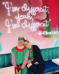Two people, dressed as elves, sit happily on a teal bench against a colorful wall with bright neon text. This joyful moment highlights caregiver support within the disability community, reflecting the mission of the Christopher Smith Foundation.| A Non Profit For Caregivers | Christopher Smith Foundation