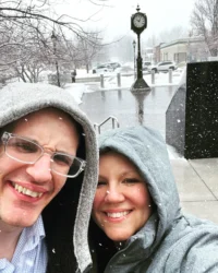 Two people in hooded coats smile in a snowy urban setting with a clock tower. This image reflects the Christopher Smith Foundation's mission of providing caregiver support within the disability community.| A Non Profit For Caregivers | Christopher Smith Foundation