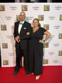 A man in a black suit with a blue bow tie stands arm in arm with a woman in a black dress on a red carpet. They smile in front of a backdrop with "Best Buddies" logos, highlighting caregiver support within the disability community.| A Non Profit For Caregivers | Christopher Smith Foundation