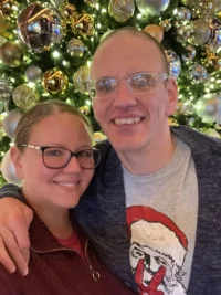 A smiling pair stands in front of a festive Christmas tree adorned with gold and silver ornaments, representing the Christopher Smith Foundation's commitment to caregivers of people with disabilities during the holiday season.| A Non Profit For Caregivers | Christopher Smith Foundation