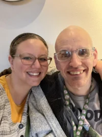 A smiling woman and man with glasses sit closely, exuding warmth and connection. The Christopher Smith Foundation highlights caregiver support, focusing on caregivers of people…| A Non Profit For Caregivers | Christopher Smith Foundation