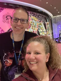 Two smiling people, one wearing glasses, stand in front of colorful art. They represent the spirit of caregiver support, embodying the Christopher Smith Foundation's mission to honor family caregivers and their dedication to the disability community.| A Non Profit For Caregivers | Christopher Smith Foundation