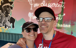 A smiling couple in casual attire poses in front of a colorful mural. The Christopher Smith Foundation supports caregivers of people with disabilities, highlighting moments of joy and connection in the disability community.| A Non Profit For Caregivers | Christopher Smith Foundation