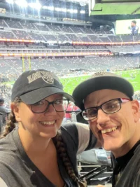 Two people wearing glasses and caps smile at a sports stadium, highlighting caregiver support within the disability community. The Christopher Smith Foundation emphasizes the importance of recognizing caregivers of people with disabilities.| A Non Profit For Caregivers | Christopher Smith Foundation
