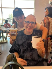A joyful moment at a cafe, showing a smiling man in glasses holding a drink, embraced by a woman in a cap. This scene highlights caregiver support and the community spirit of the Christopher Smith Foundation.| A Non Profit For Caregivers | Christopher Smith Foundation