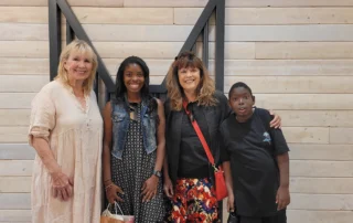 Four smiling people stand together in front of a light wooden wall, embodying the spirit of caregiver support celebrated by the Christopher Smith Foundation.| A Non Profit For Caregivers | Christopher Smith Foundation