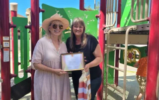 Two women stand in a playground, smiling and holding a framed certificate from the Christopher Smith Foundation, celebrating caregiver recognition for their dedication to supporting the disability community.| A Non Profit For Caregivers | Christopher Smith Foundation
