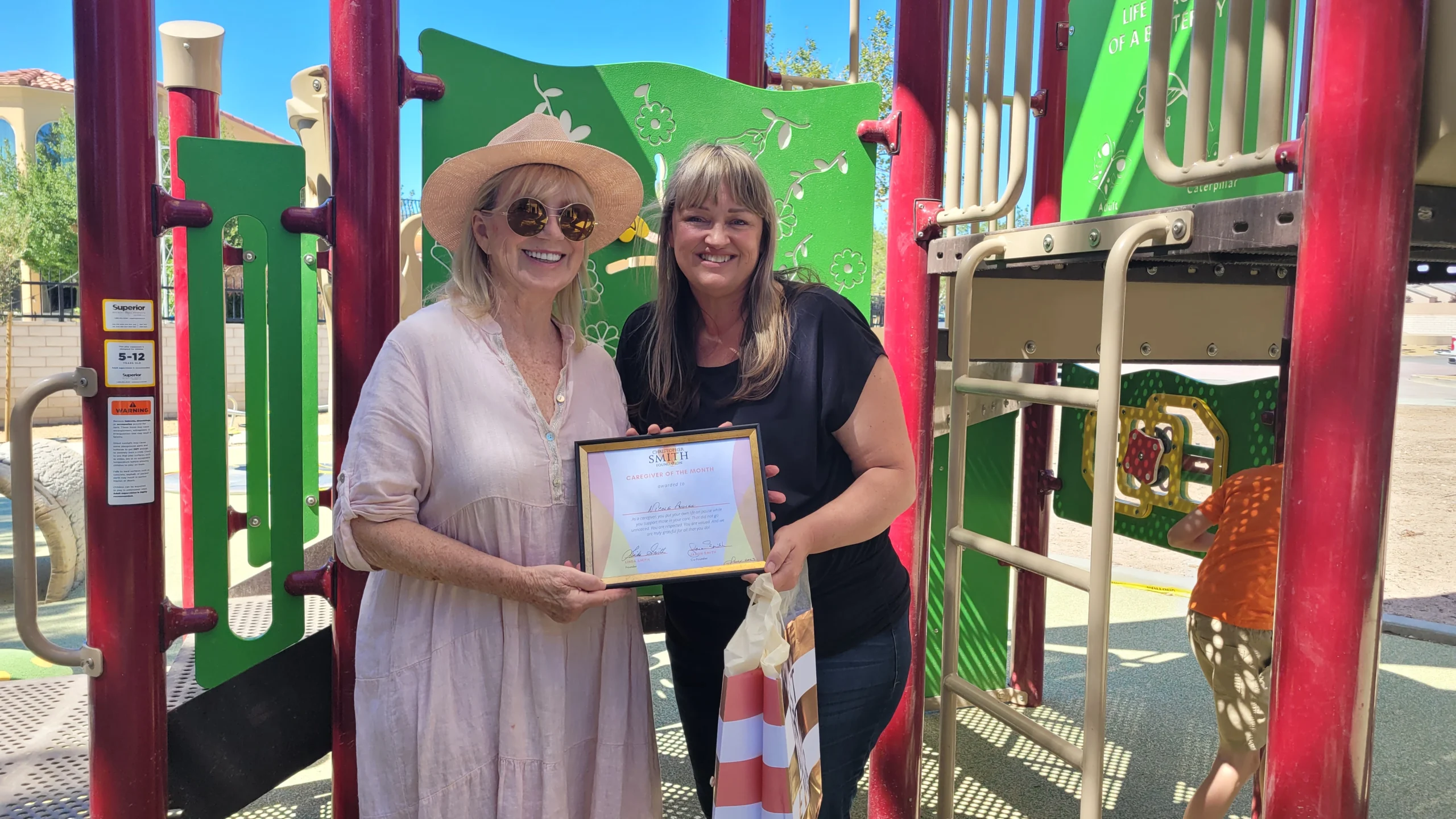Two women stand in a playground, smiling and holding a framed certificate from the Christopher Smith Foundation, celebrating caregiver recognition for their dedication to supporting the disability community.| A Non Profit For Caregivers | Christopher Smith Foundation
