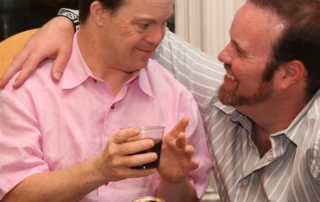 A man in a pink shirt smiles warmly at another man in a striped shirt, both holding drinks, embodying family caregivers' support, a core mission of the Christopher Smith Foundation.| A Non Profit For Caregivers | Christopher Smith Foundation