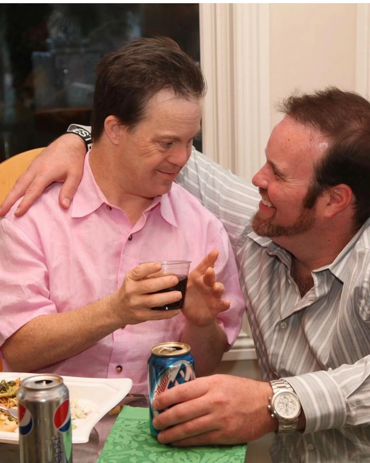A man in a pink shirt smiles warmly at another man in a striped shirt, both holding drinks, embodying family caregivers' support, a core mission of the Christopher Smith Foundation.| A Non Profit For Caregivers | Christopher Smith Foundation