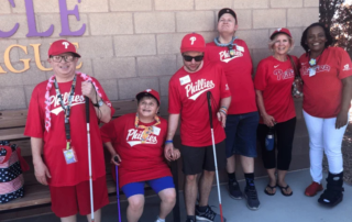 A group of people wearing matching red Phillies shirts and caps, some holding white canes, stands smiling in front of a brick wall. This image represents the Christopher Smith Foundation's caregiver support and advocacy for the disability community.| A Non Profit For Caregivers | Christopher Smith Foundation