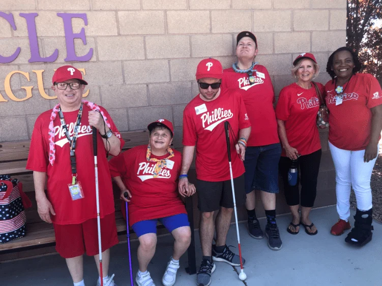 A group of people wearing matching red Phillies shirts and caps, some holding white canes, stands smiling in front of a brick wall. This image represents the Christopher Smith Foundation's caregiver support and advocacy for the disability community.| A Non Profit For Caregivers | Christopher Smith Foundation
