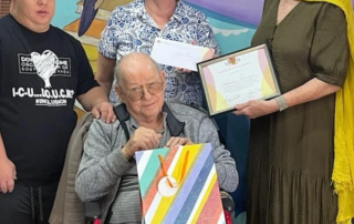 A group of people, including a person in a wheelchair, smile while holding a colorful gift bag and certificate, celebrating caregiver support at a Christopher Smith Foundation event.| A Non Profit For Caregivers | Christopher Smith Foundation