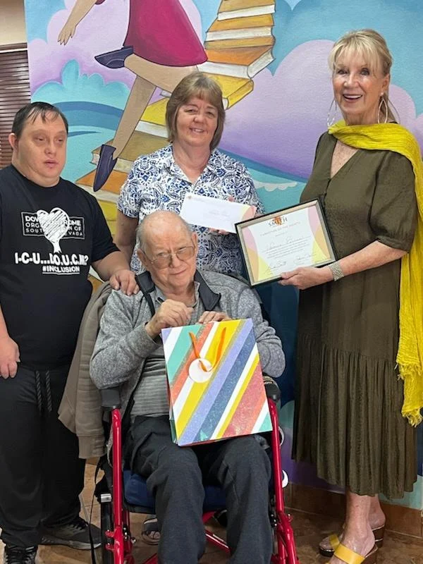 A group of people, including a person in a wheelchair, smile while holding a colorful gift bag and certificate, celebrating caregiver support at a Christopher Smith Foundation event.| A Non Profit For Caregivers | Christopher Smith Foundation